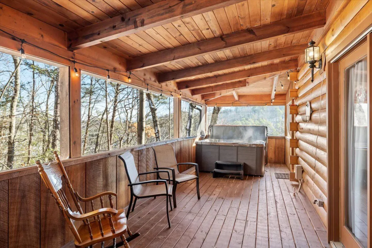 An inviting outdoor hot tub sits on a wooden deck, accompanied by a classic rocking chair. The surrounding beams and large windows frame lush forest views, suggesting privacy and tranquility. The ambiance hints at both daytime relaxation and romantic stargazing at night.