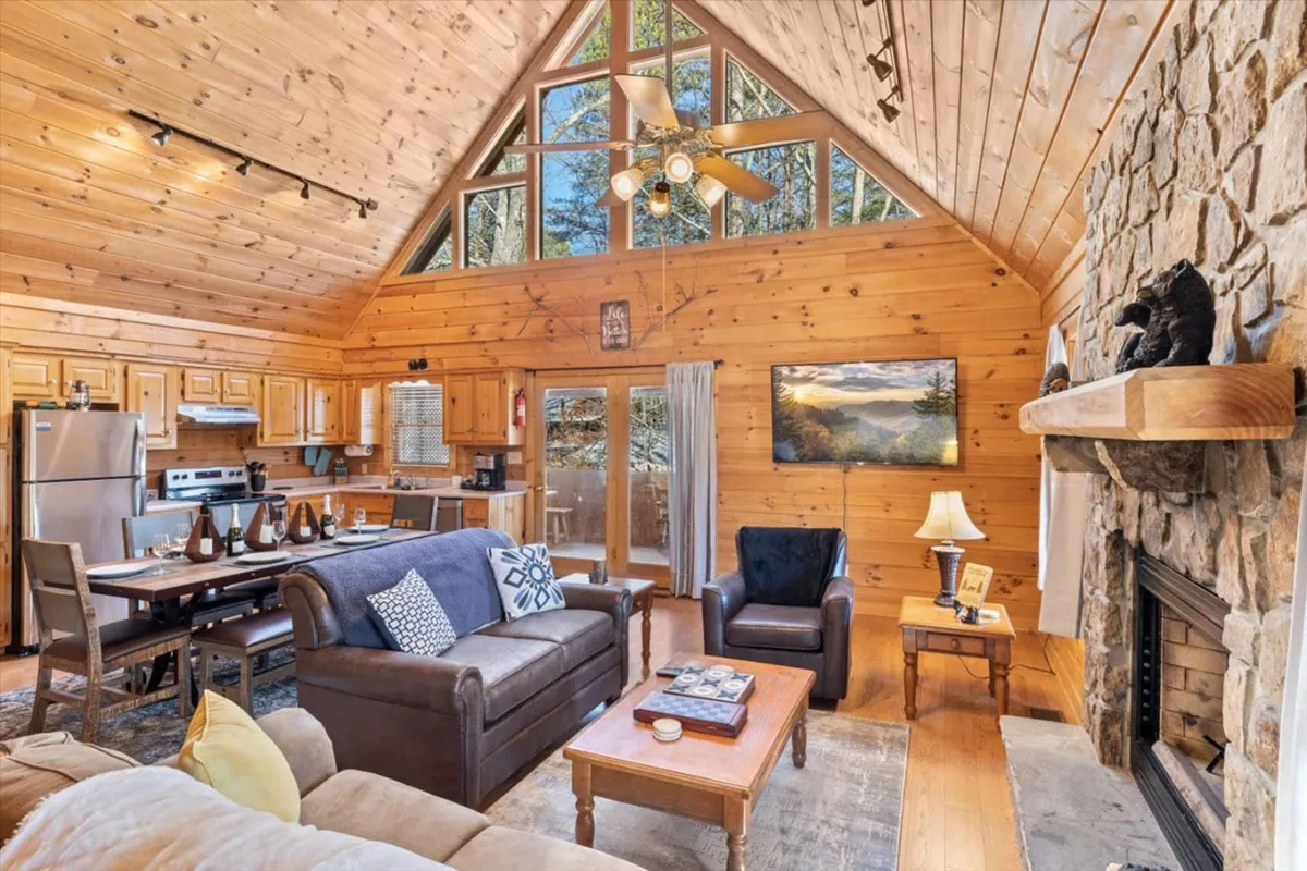 Inside, a warm open-concept living area features a striking stone fireplace, exposed wooden beams, expansive windows that let in natural light, and a spacious dining table ready for group meals. The cozy yet spacious layout is ideal for laughter, shared stories, and creating lasting memories together.