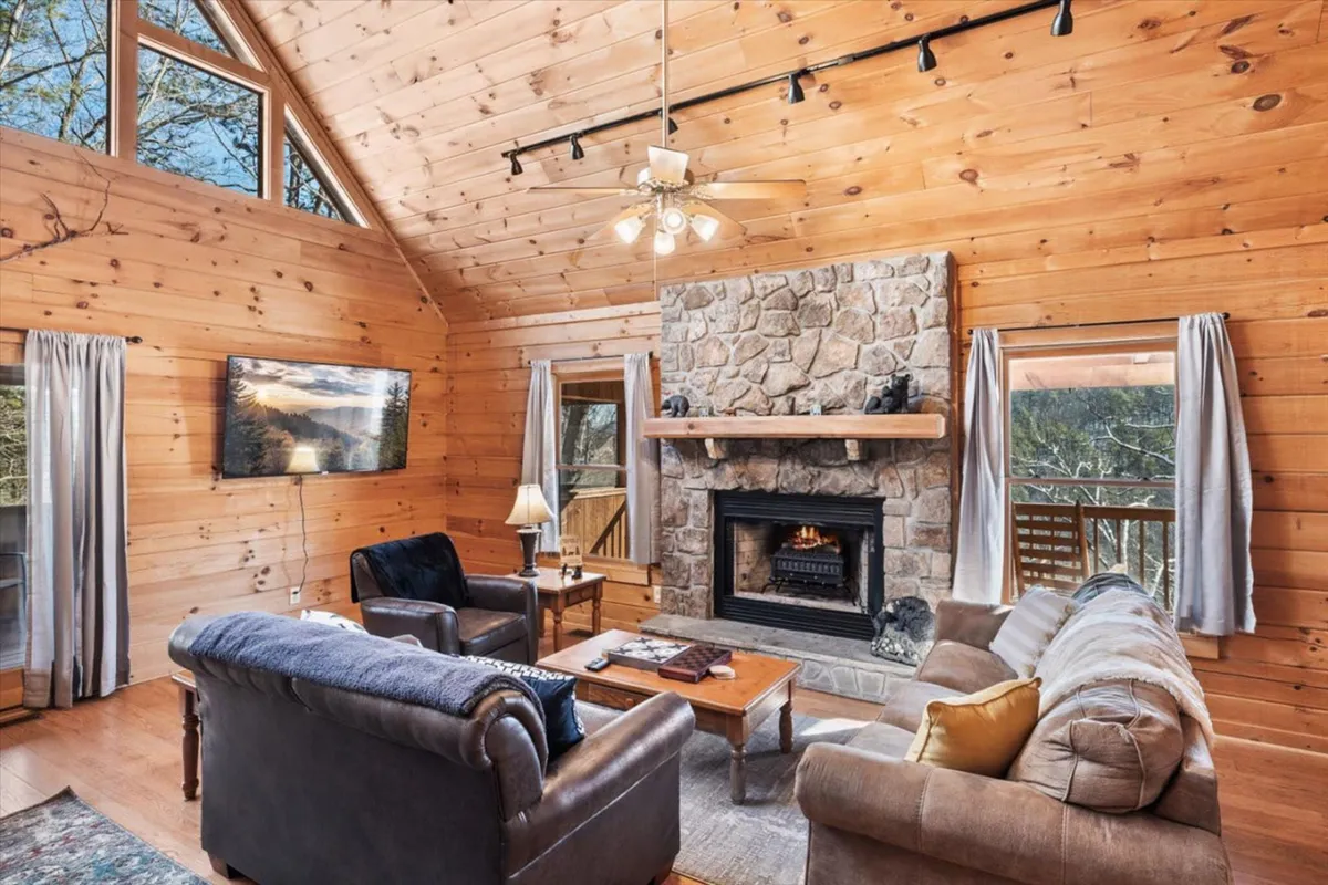 A cozy living area with a stone fireplace as its centerpiece, comfortable seating, large windows for natural light, and a wooden ceiling. The scene is set for relaxation, socializing, or gathering by the fire for a movie night.