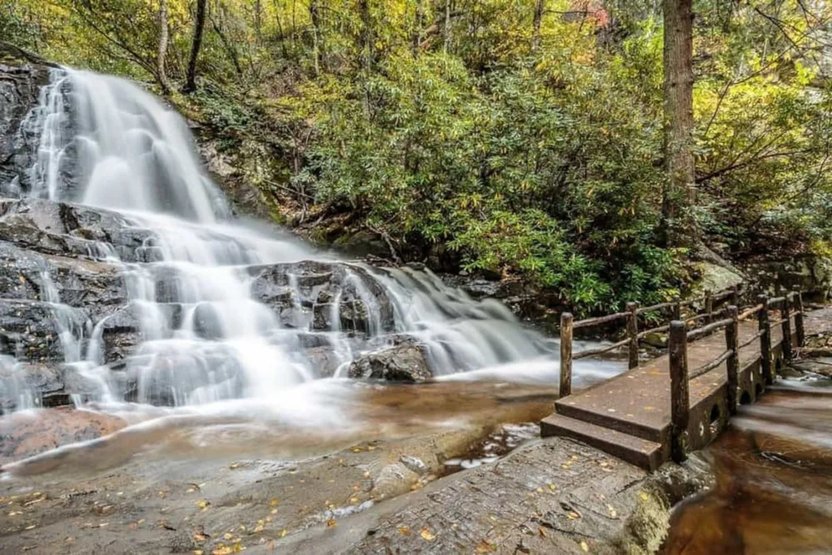 A picturesque waterfall surrounded by lush foliage, with a wooden bridge offering a scenic vantage point. The setting is ideal for hiking, nature watching, and enjoying the serenity of the Smoky Mountains region.