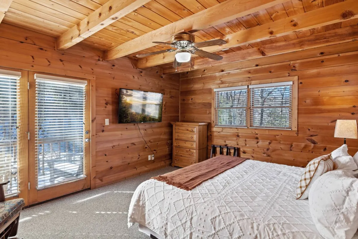 A warmly lit king bedroom featuring a large, plush bed with crisp linens, a wooden ceiling, dresser, and a wall-mounted television. The patio door opens onto a private balcony, letting in natural light and offering a glimpse of the outdoors.