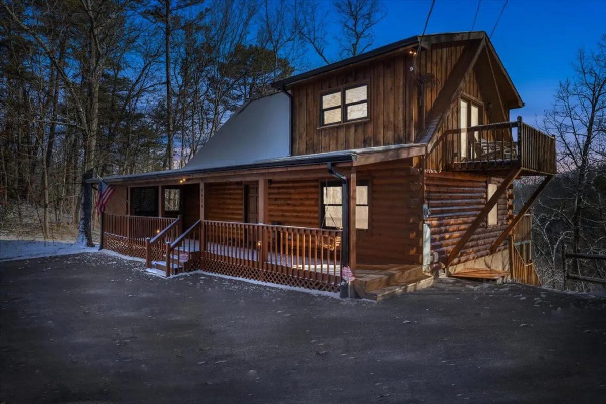 An exterior view of the cabin’s wooden structure with a balcony, outdoor stairs, and an American flag. The yard and surrounding trees set a tranquil, mountain atmosphere, perfect for outdoor gatherings or quiet moments reflecting in nature.