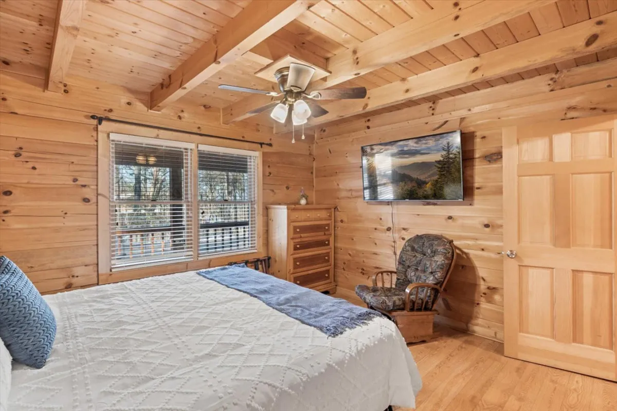 A tranquil bedroom with a king-size bed dressed in blue accents, wooden ceiling and walls, a TV displaying a scenic image, and a cozy armchair placed by the window. Sunlight streams in, creating a serene and restful atmosphere.