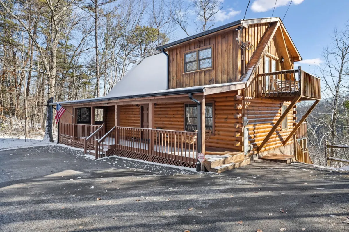 A charming log cabin exterior with a welcoming front porch, wooden balcony, and an American flag. The setting suggests a cozy home base for families, perfect for relaxing after outings and enjoying the fresh mountain air.