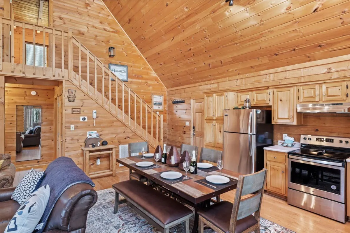 A warm, inviting open-concept space featuring a wooden dining table set for a group, a staircase leading to another level, and a glimpse of the kitchen with modern appliances. The area is surrounded by wooden walls, enhancing the cozy ambiance and highlighting the seamless flow between dining, kitchen, and living spaces.