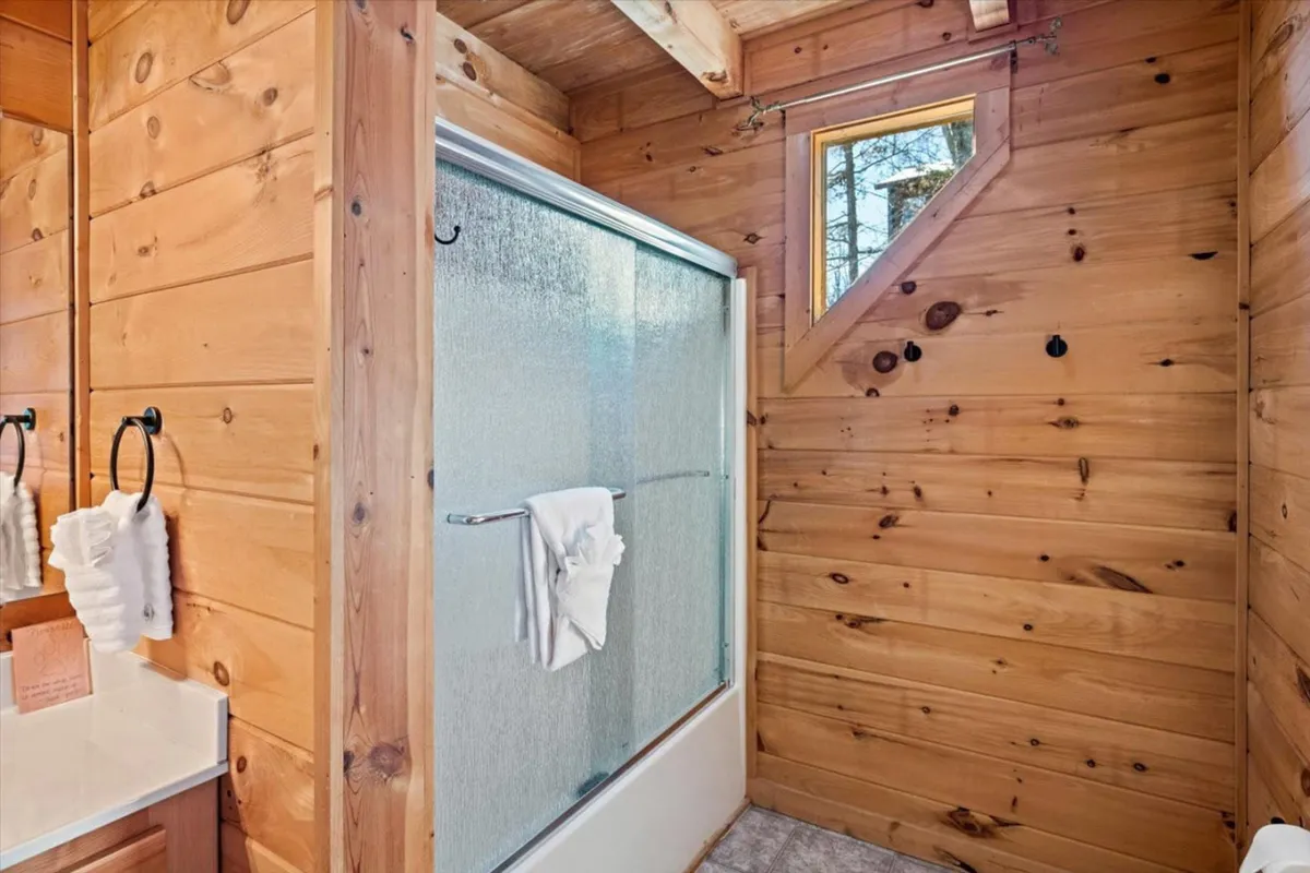A modern ensuite bathroom with a glass shower enclosure, wood paneling, and a small window letting in natural light. The space is clean, inviting, and designed for relaxation.