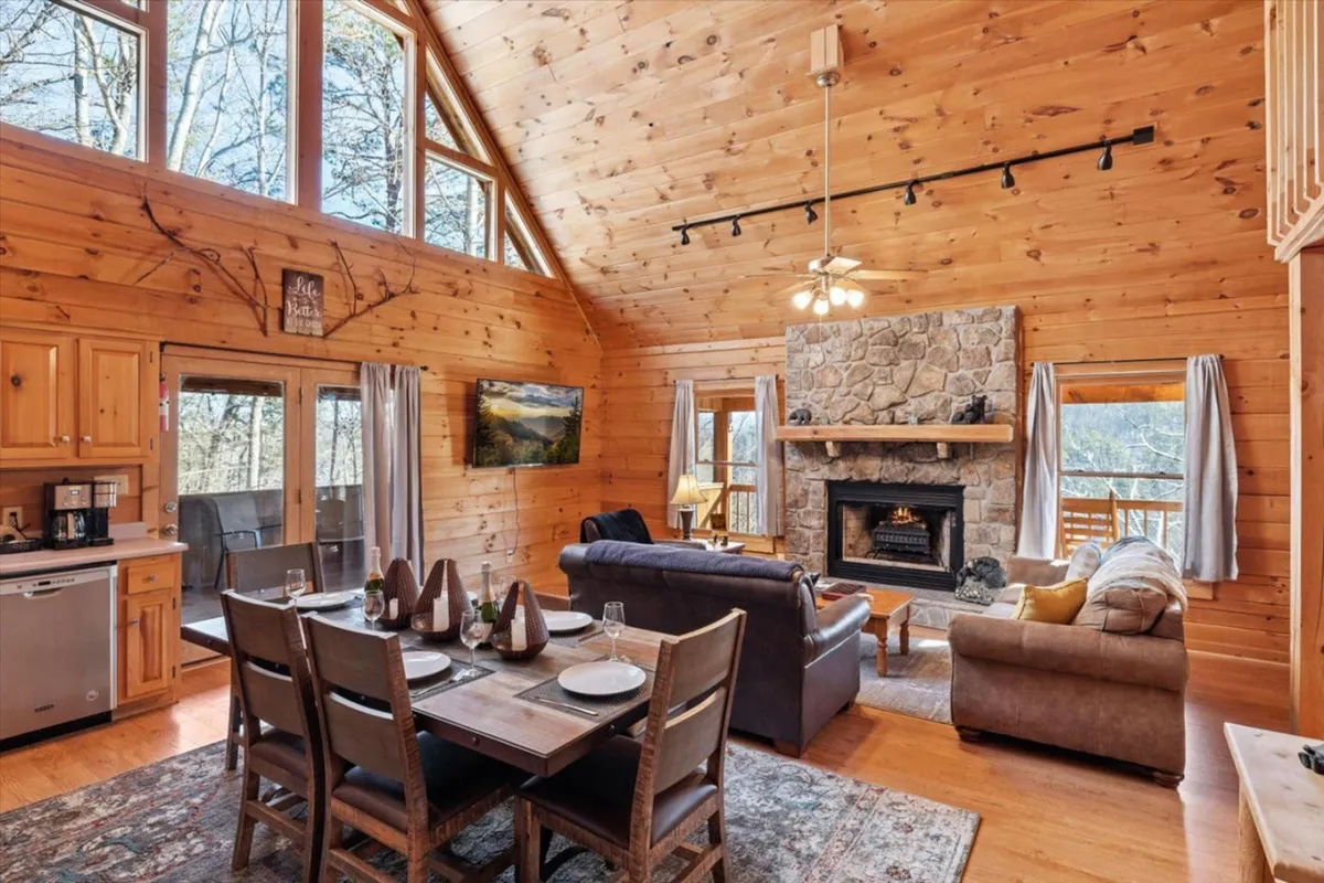 The image showcases the inviting interior of a mountain log cabin, centered around a rustic stone fireplace, with expansive windows framing lush forest views. A spacious dining table sits nearby, ideal for family gatherings or holiday meals. The atmosphere is cozy and serene, perfect for relaxing evenings after exploring Sevierville's scenic surroundings.